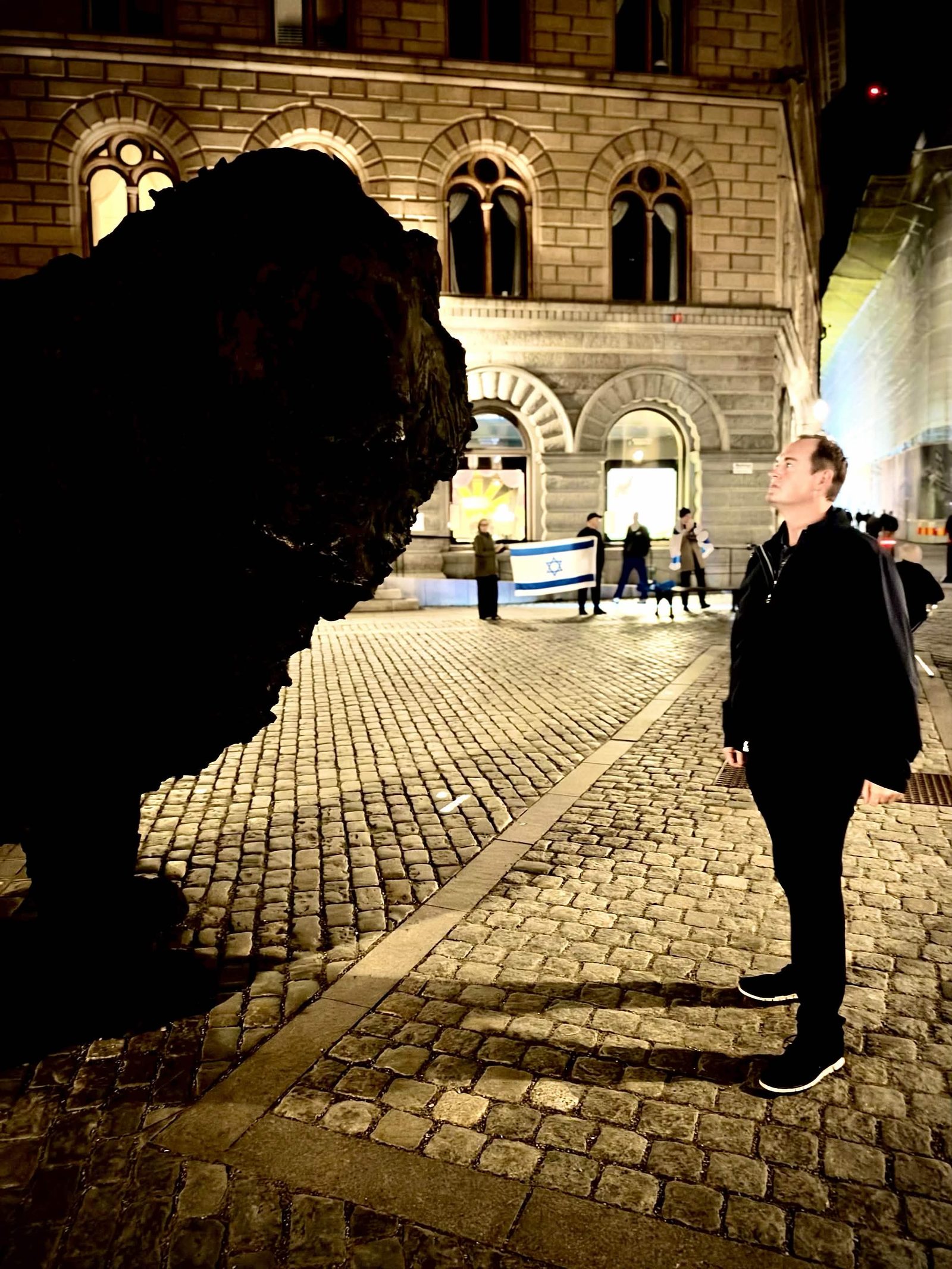 A night plaza on the trip. A darker lion. A lit stone front. An Israeli flag in the background. The city is not named.
